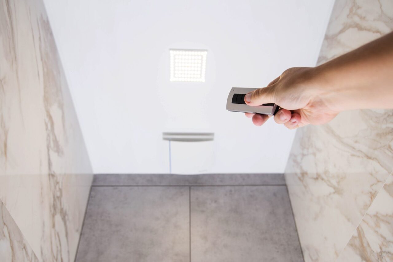 Personne réglant un capteur d’humidité près d’une bouche de VMC dans une salle de bains sans fenêtre.